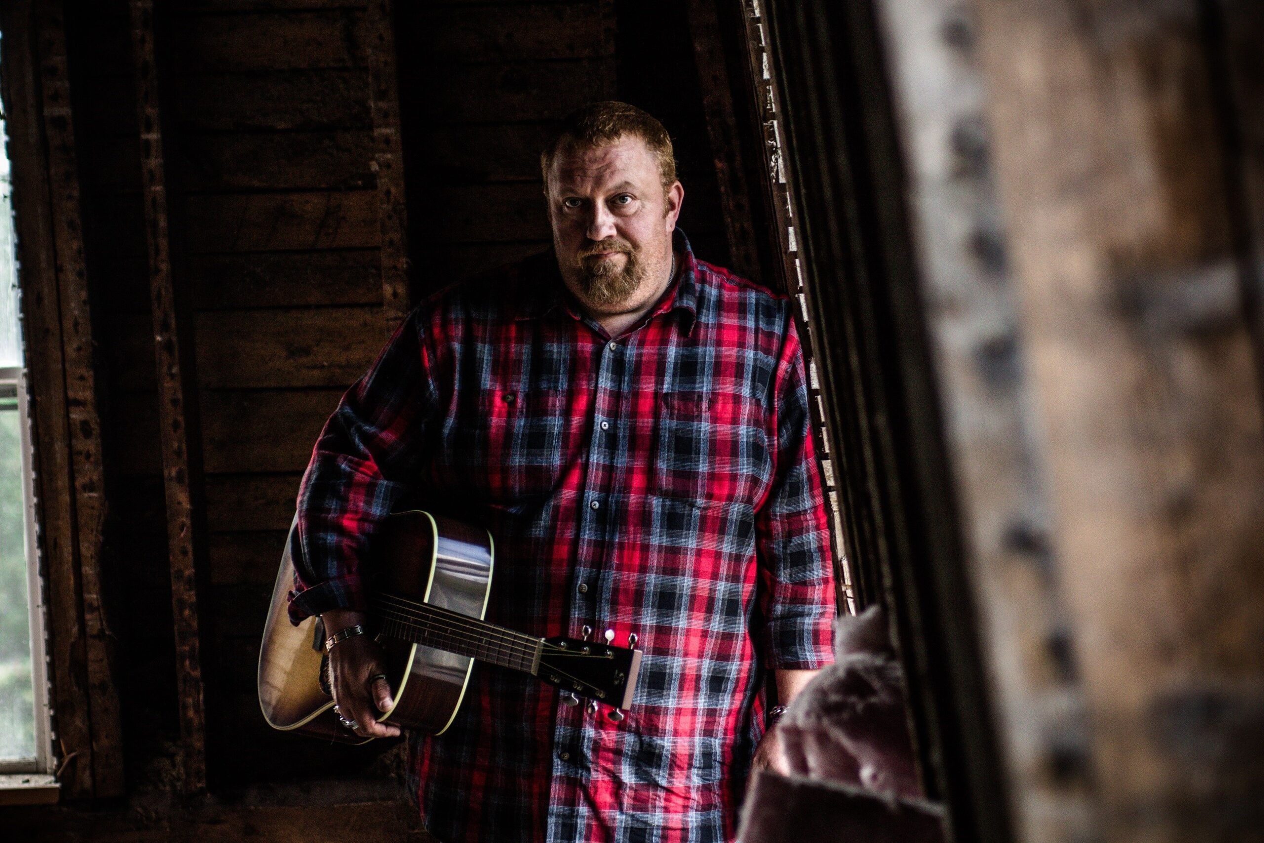 J.P. Cormier wearing a read plaid shirt with guitar under his arm. Background is an old barn loft